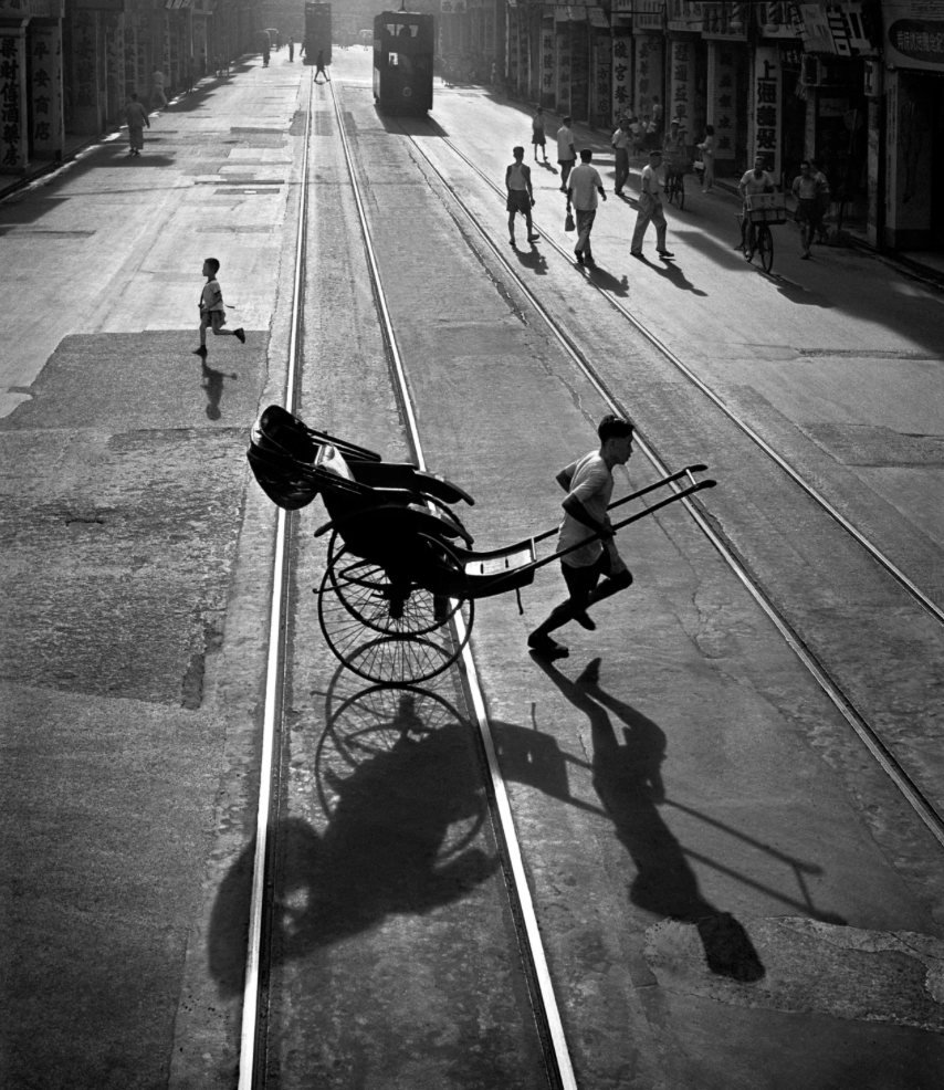 Fotografia de Fan Ho mostrando um puxador de riquixá correndo sobre trilhos de bonde em Hong Kong